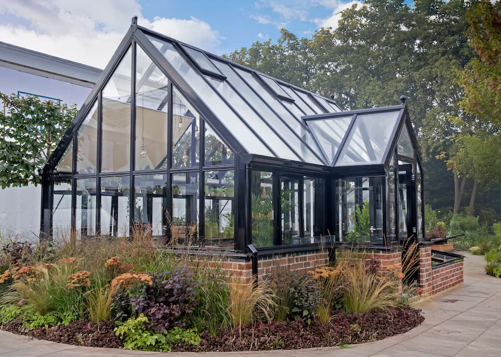 A large aluminium framed greenhouse in black on a brick base with season planting around it
