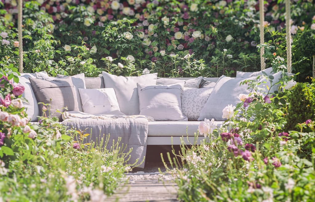 A padded swinging sofa in the garden with an abundance of soft cushions in cream and white. Roses and other plants surround.