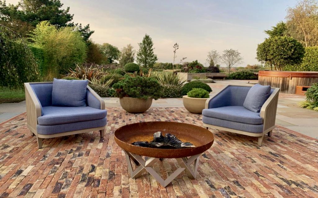 Contemporary chairs with fire pit in large rural garden with clay pavers and evergreen planting in the backgound