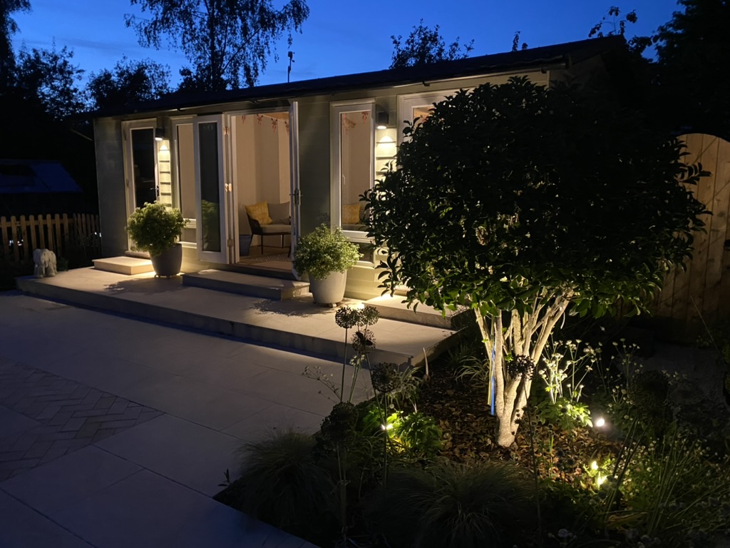 Soft lighting uplights a multi-stem tree near a terrace. A garden room is in the back ground. The lighting creates a cosy look.