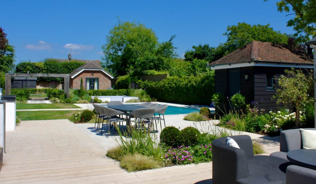 A wide angle view of a contemporary garden which shows lush planting interspersed with different hard landscaping. Paving is porcelain so smooth and contemporary. In the mid ground is a swimming pool which is next to a lawn which allows children to play with ease