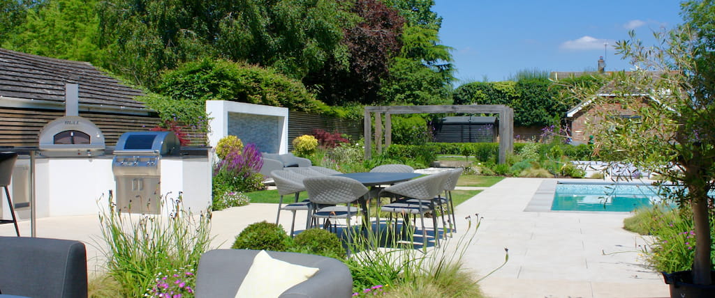 A high end luxury garden with contemporary features including outdoor kitchen with Bull grill and Bull pizza oven. Stylish planting breaks up the hard landscaping, nearby a swimming pool has been designed to ensure this fun entertaining garden is arranged to ensure it it suitable for a family. Oversized pergolas create a statement to the rear and frames the rear section of the garden. Luxury garden by Llevelo Garden Design.