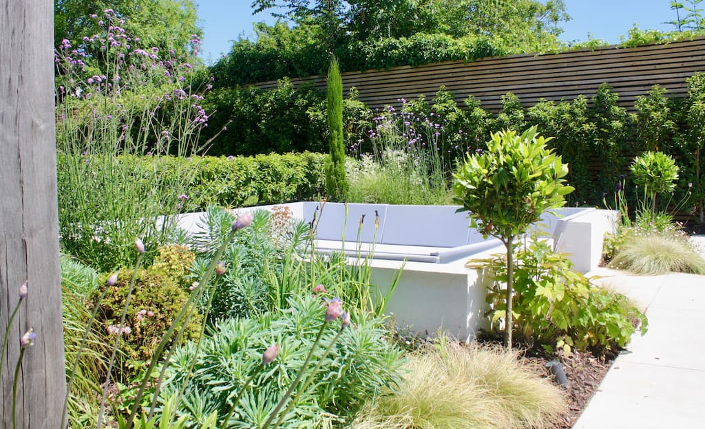 A luxury and stylish sunken seating area with central fire pit. Modern slatted fencing to the rear, evergreen planting surrounds the seating area and provides a feel of cosiness. The seating is completed in stylish greys. A muted colour palette gives the space an elegant and contemporary finish.