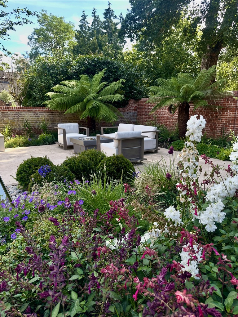 A lush garden corner or a high end walled garden in Essex. Two tree ferns flank a seating area with tables surrounding a fire table. Colourful planting in the foreground is abundant.