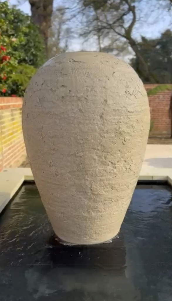 A close up of a taupe coloured concrete urn which is textured. Water trickles down the sides. The urn sits centrally to a water feature in a walled garden
