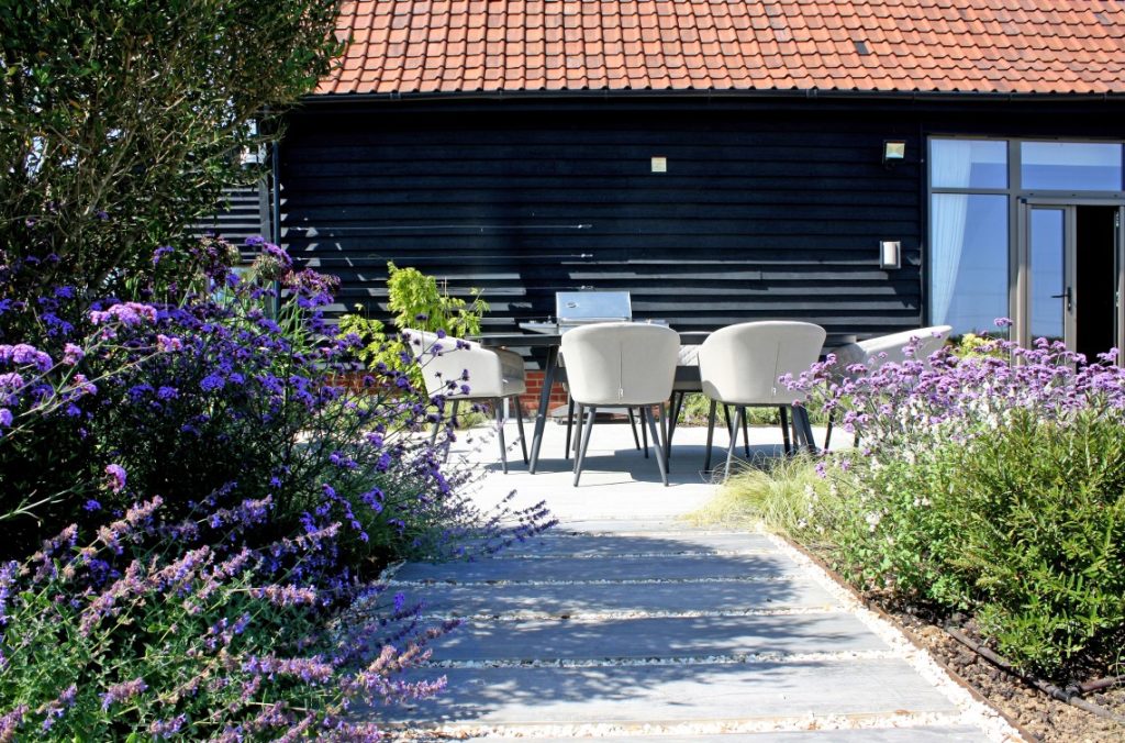 Modern patio furniture in the distance with cottage style planting and a tree in the foreground. A stepping stone style path with pavers in gravel leads the eye to the furniture setting in the mid ground. A black barn conversion is in the background.