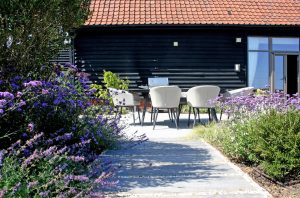 Modern patio furniture in the distance with cottage style planting and a tree in the foreground. A stepping stone style path with pavers in gravel leads the eye to the furniture setting in the mid ground. A black barn conversion is in the background.