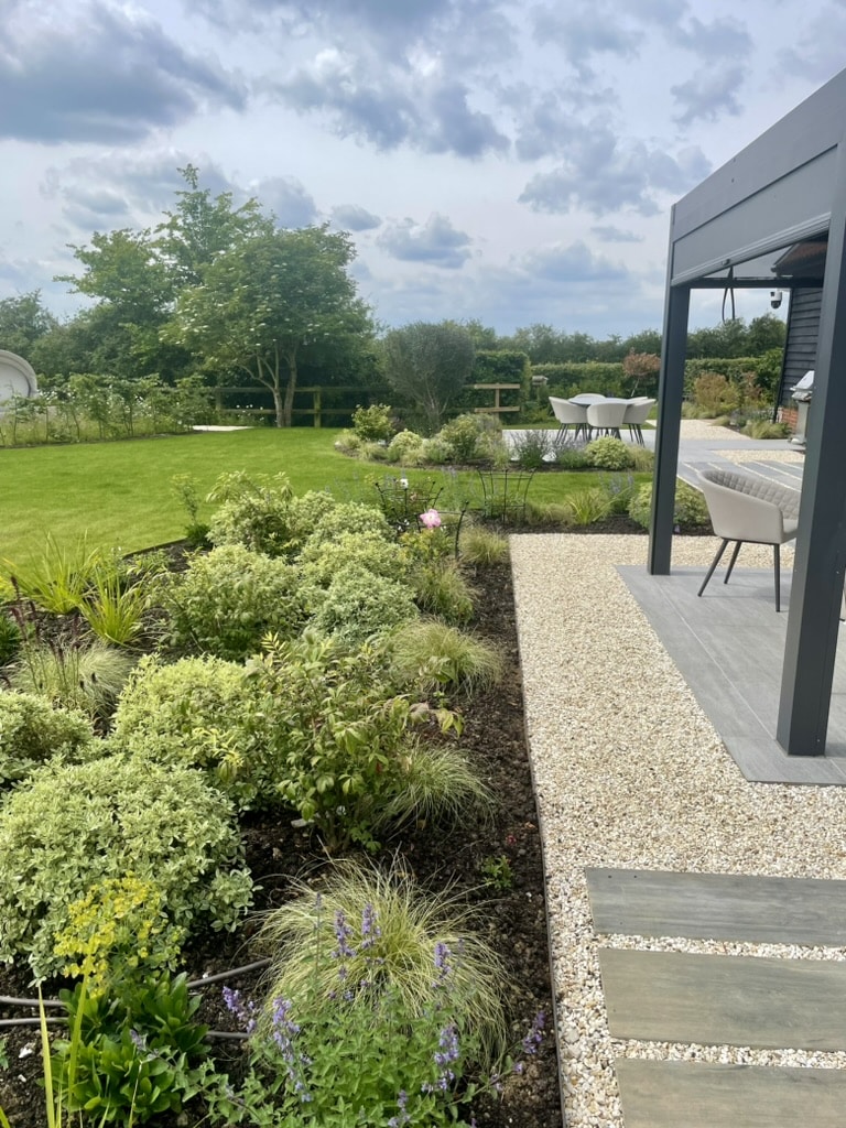 A view across a garden. To the right, the edge of a pergola can be seen. There is a gravel walkway next to this and a planting border with green shrubs and perennials to the left of that. In the distance is dining area and more planting. Lawn is the midground.