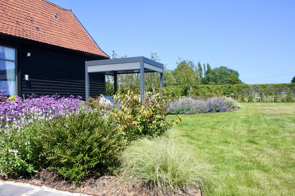 Garden scene for a converted barn. The barn can be seen to the left of the image, a modern grey pergola. Modern planting with green and purple tones can be seen in the foreground.