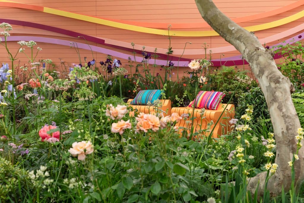 A garden scene that includes a brightly coloured rear boundary with thin panels of coloured timber waving horizontally across the image. Two orange rocking seats are positioned in the centre of the garden and bright and colourful planting is in the foreground.