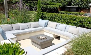 Image shows a sunken fitted seating area in a garden. Seats are dressed with fitted and decorative cushions and form a square around a modern fire table